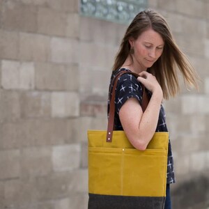 May include: A woman is holding a yellow and black canvas tote bag with brown leather straps. The bag has a large pocket on the front.