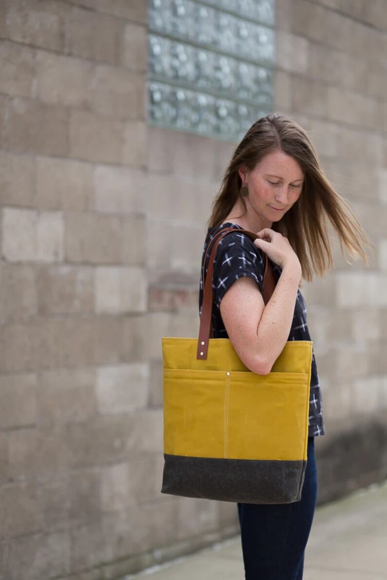 May include: A woman is holding a yellow and black canvas tote bag with brown leather straps. The bag has a large pocket on the front.