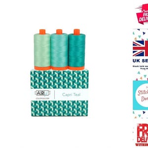 Puede incluir: Tres carretes de hilo de coser en varios tonos de azul verdoso, desde menta claro hasta turquesa intenso, están dispuestos en fila sobre una caja rectangular. La caja presenta un patrón geométrico y el texto Aurifil Color Builder Capri Teal.