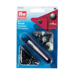 May include: A package of 10 black 'Anorak' press fasteners, also known as snap buttons, with a diameter of 15 mm. The package includes a tool for attaching the fasteners. The package is blue and white with the Prym logo and the text 'Anorak' and 'Press fasteners'.