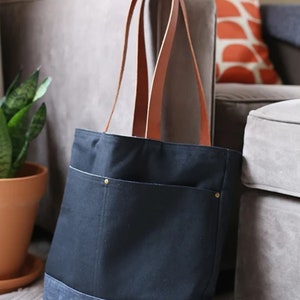 May include: A black canvas tote bag with a denim bottom and brown leather straps. The bag has two front pockets with brass rivets.