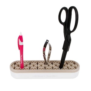 May include: A beige and white organizer with a geometric pattern holds a pink pen, a pair of scissors, and a small metal tool. The organizer is made of silicone and has a rectangular shape.