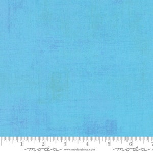 May include: A solid blue fabric with a subtle textured pattern. The fabric is a light blue colour with a slightly faded look. The fabric is marked with a ruler and the word "moda" in a handwritten font.