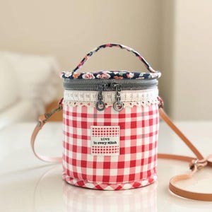 Peut inclure: Sac cylindrique en tissu vichy rouge et blanc, avec une poignée fleurie et une bandoulière en cuir marron. Le sac est orné d'une bordure en dentelle blanche et d'une étiquette "Love in every stitch".