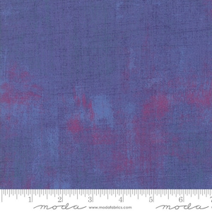 May include: A close-up of a blue fabric with a subtle pink and blue mottled pattern. The fabric has a textured surface and a slight sheen. The fabric is marked with a ruler in inches and the word "moda" is printed on the ruler.