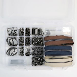 Könnte beinhalten: Eine transparente Kunststoffbox mit verschiedenen Näh- und Bastelmaterialien. Die Fächer enthalten Metallschnallen, Nieten, Ringe, Reißverschlüsse in Braun, Marineblau und Creme sowie eine Kette. Die Hardware ist hauptsächlich schwarz.