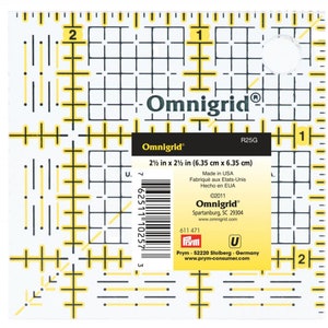May include: A clear Omnigrid R25G quilting ruler, measuring 6.35 cm x 6.35 cm, with black and yellow grid lines and measurements. The ruler has the Omnigrid logo and product information printed on it. Made in the USA.