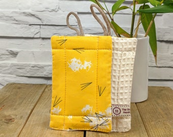May include: A yellow dishcloth with a white and black arrow pattern and a white waffle weave dishcloth with a brown button and the text "The Stitch Hand" on it.