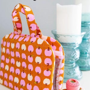 May include: An orange quilted bag with a repeating pattern of pink and white apples. The bag, called The Truvy Bag, has a clear side panel and a handle. The words "Knot + Thread Design" are in a black box on the bottom left.