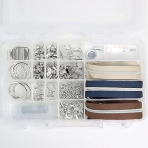 Op de afbeelding: Een doorzichtige plastic organizer gevuld met diverse naai- en knutselbenodigdheden. De vakken bevatten zilverkleurige metalen hardware, waaronder ringen, klinknagels en kettingen, samen met drie ritsen in beige, marineblauw en bruin.