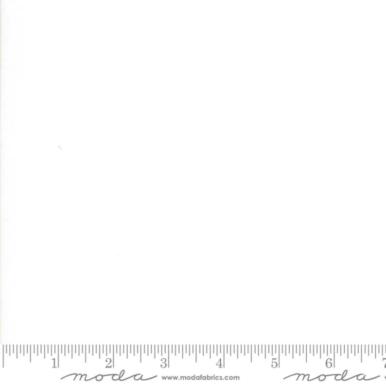 May include: A plain white fabric swatch with a ruler at the bottom. The ruler is marked in inches and has the text "moda" printed on it.