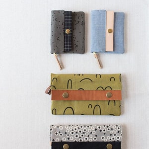 May include: Four different fabric wallets with various patterns and colors. The wallets have a flap closure with a button. The patterns include a green and black abstract design, a gray and white polka dot design, a blue denim design, and a gray and black speckled design.
