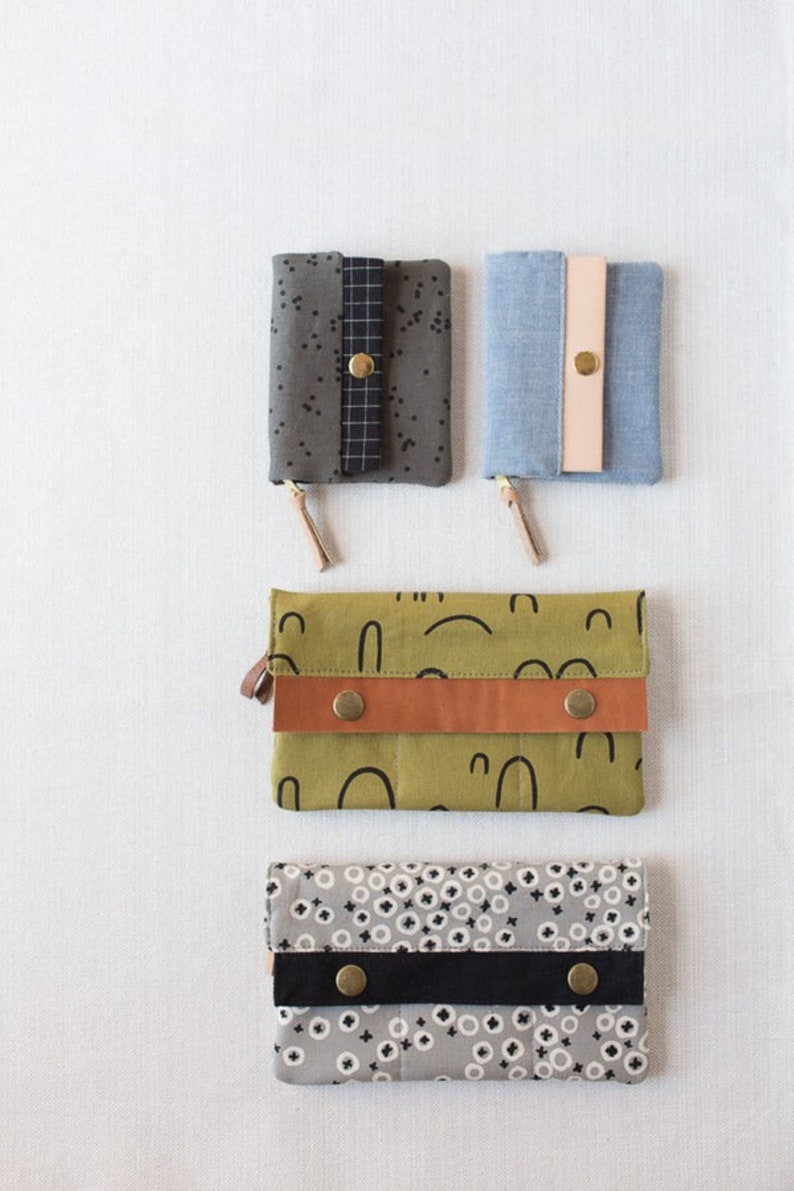 May include: Four different fabric wallets with various patterns and colors. The wallets have a flap closure with a button. The patterns include a green and black abstract design, a gray and white polka dot design, a blue denim design, and a gray and black speckled design.