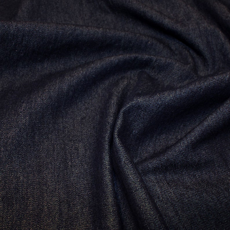 May include: Close-up of a dark blue denim fabric with a textured weave. The fabric is folded and draped, showing the soft texture and subtle sheen.
