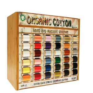 Op de afbeelding: Houten display met spoelen gekleurd garen. De display toont "Organic Cotton" en "Hand and Machine Quilting". De garenkleuren zijn wit, roze, blauw, groen, oranje en bruin. De tekst "Extra Long Staple Egyptian Cotton" is ook zichtbaar.