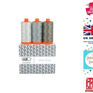 May include: Three spools of sewing thread in varying shades of grey, ranging from light to dark, are positioned on top of a rectangular box. The box features a geometric pattern of blue, white, and grey cubes and is labeled Auri Color Builder.