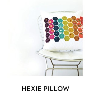 May include: A white pillow with a colourful geometric pattern of hexagons in shades of pink, orange, yellow, green, blue, and purple. The pillow is sitting on a silver chair. The text "HEXIE PILLOW" is below the image, and the text "MODERN HANDCRAFT" is below that.