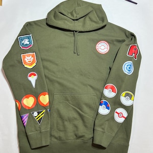 May include: A green hooded sweatshirt with various colorful patches and embroidered designs. The patches include a Pokeball, a Pokemon character, and a heart.