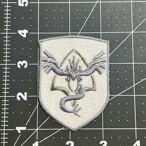 May include: Embroidered patch with a shield shape, featuring a stylized design of a mythical creature with wings and a serpentine body. The patch is primarily light gray with gray and tan accents.