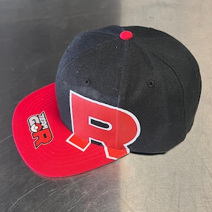 May include: Black and red baseball cap with a flat brim. The cap features a large red "R" on the side and the text "Team Go R" on the brim. A red button is on top.