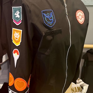 May include: A black bomber jacket featuring a silver front zip and a stand-up collar. The right sleeve is adorned with a vertical row of colourful embroidered patches, while the chest displays a blue bird emblem and a circular red and white logo.