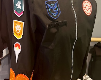 May include: A black bomber jacket featuring a silver front zipper and a stand-up collar. The right sleeve is adorned with a vertical row of colorful embroidered patches, while the chest displays a blue bird emblem and a circular red and white logo.