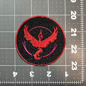May include: A round, black embroidered patch featuring a red phoenix design within a pink circle. The phoenix has outstretched wings and a flame. The patch is likely for clothing or accessories.