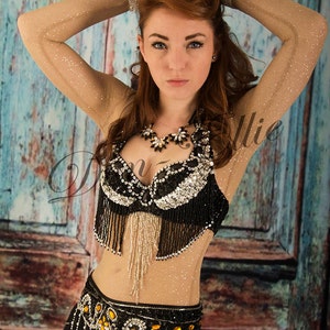 May include: A woman wearing a black and silver beaded belly dancing costume with a fringe bra and a belt with gold accents. She is posing with her arms raised above her head.