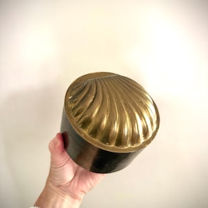Peut inclure: Une boîte ronde de couleur laiton avec un couvercle en forme de coquillage. Le couvercle présente un motif détaillé et nervuré. La boîte est tenue dans une main sur un fond uni blanc cassé. La boîte semble être en métal.