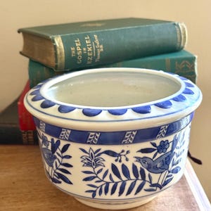 May include: A white ceramic bowl featuring a blue painted pattern of birds and foliage. The rim has a scalloped blue edge, and the body displays a repeating botanical design with small birds. The bowl has a wide opening and a rounded base.