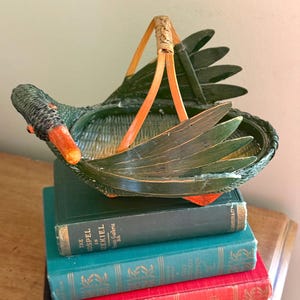 May include: A dark green woven basket shaped like a swan with an orange beak and eyes. The basket features a light brown arched handle and stylized wing-shaped sides. The body is constructed from woven material with a flat base and a central opening.