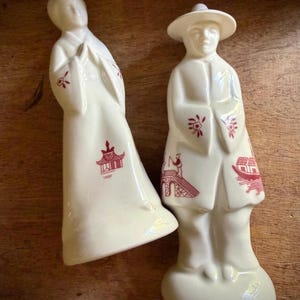 May include: Two cream-coloured ceramic figurines. One is a woman in a long robe, the other a man in a hat and coat. Both have red decorative accents, including pagodas and bridges. The figurines are likely decorative items or collectibles.