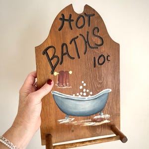 May include: Wooden sign with the words "HOT BATHS 10¢" in black script. The sign features a painted blue bathtub with bubbles, a towel rack, and a brown towel. The sign is held by a person.