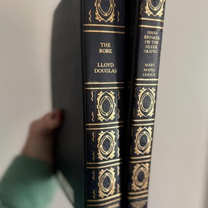May include: Two hardcover books with black covers and gold lettering. The book on the left is titled "The Robe" by Lloyd Douglas. The book on the right is titled "Hans Brinker or the Silver Skates" by Mary Mapes Dodge. Both books have decorative gold accents.