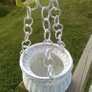 May include: A white wicker hanging basket with a round shape and a woven design. The basket is suspended by white chain links. The background features green grass and a wooden post.