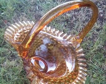May include: Amber-colored glass basket with a handle. The basket has a ruffled edge and a textured, dimpled surface. The handle arches gracefully over the bowl. The basket is sitting on a bed of green grass.