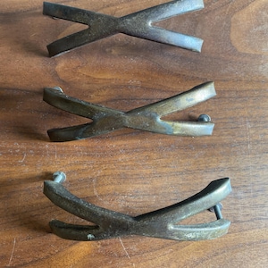 May include: Three antique brass-toned cabinet pulls in an X-shape. The pulls are slightly curved and have a vintage appearance. Each pull has a screw for mounting. The pulls are arranged on a wooden surface.