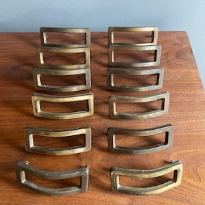 May include: A collection of twelve vintage brass drawer pulls. Each pull has a rectangular shape with a curved handle. The brass has a warm, aged patina. The pulls are arranged on a wooden surface.