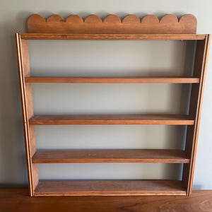 May include: A medium-brown wooden wall shelf featuring four horizontal tiers for storage. The top edge of the unit is finished with a decorative scalloped trim. The structure is composed of vertical side panels and four flat shelves.