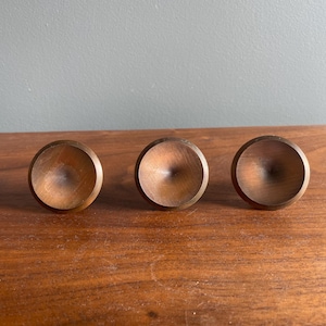 Puede incluir: Tres pomos de madera con un diseño redondeado en forma de cuenco. Los pomos son de color marrón cálido y parecen estar hechos de madera maciza. Están dispuestos en línea horizontal sobre una superficie de madera.
