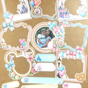 May include: A collection of die-cut paper frames and labels featuring illustrations of castles, ferris wheels, balloons, bows, and flowers. One circular frame contains a photograph of two people. The designs include pastel colors and park-themed icons.
