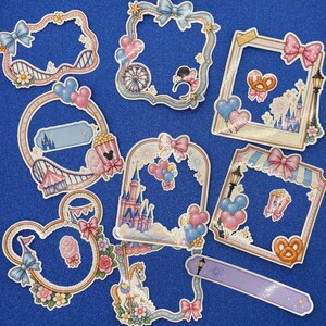 May include: A collection of die-cut stickers featuring various decorative frames, including shapes with bows, amusement park elements like a carousel horse, a Ferris wheel, a castle, popcorn, pretzels, and balloons in shades of pink, blue, and gold.