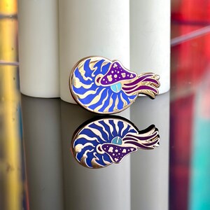 Op de afbeelding: Een gouden emaille pin met een nautilus schelp met blauwe en witte strepen en een paarse en roze kop met een blauw oog. De pin is een grillige en kleurrijke weergave van het zeedier.