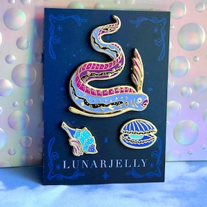 Puede incluir: Un conjunto de tres pines de esmalte dorado con una serpiente marina púrpura y azul, una concha de mar azul y una concha de almeja azul. Los pines están sobre un fondo negro con el texto "LUNARJELLY" en dorado.