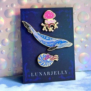 Op de afbeelding: Een set van drie emaillepins met een blauwe walvis, een nautilus schelp met een paddenstoel en een roze paddenstoel. De pins zijn op een donkerblauwe kaart met de tekst "LUNARJELLY".