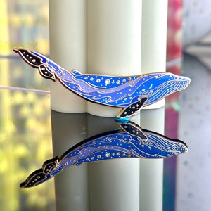 Op de afbeelding: Een blauwe en paarse emaille pin van een walvis met een sterrenhemelontwerp. De walvis kijkt naar rechts en heeft een gedetailleerde omtrek van zijn lichaam. De pin wordt weerspiegeld in een spiegelachtig oppervlak.