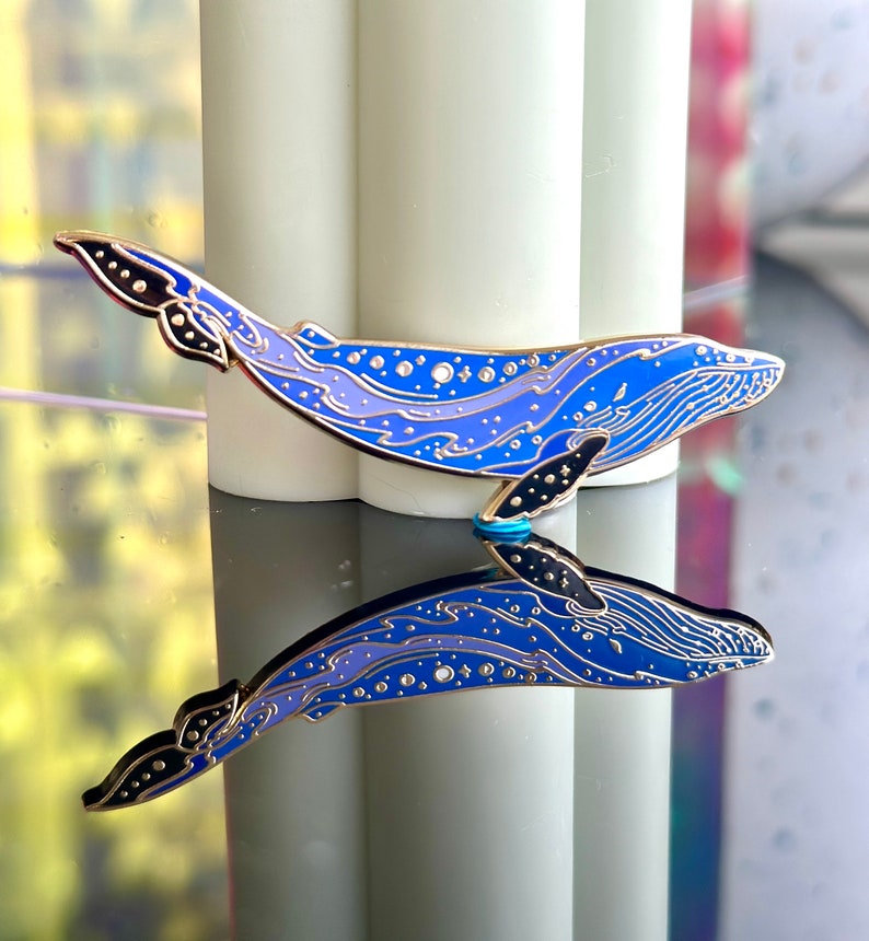 Op de afbeelding: Een blauwe en paarse emaille pin van een walvis met een sterrenhemelontwerp. De walvis kijkt naar rechts en heeft een gedetailleerde omtrek van zijn lichaam. De pin wordt weerspiegeld in een spiegelachtig oppervlak.