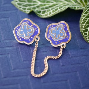 May include: A pair of gold-tone metal flower-shaped brooches with blue and light blue enamel. The brooches are connected by a gold chain.