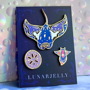 Op de afbeelding: Een set van drie emaillepins met een blauwe mantarog, een paarse zeekomkommer en een paarse en roze zeester. De pins zijn op een zwarte achtergrond met gouden accenten en de tekst "LUNARJELLY".