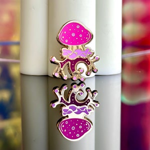 Op de afbeelding: Een gouden emaille pin met een roze en paarse paddenstoel met een draaiende steel. De paddenstoel heeft witte stippen en een gouden omtrek.